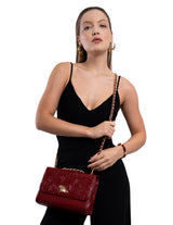 A woman wearing a black spaghetti-strap dress models a deep red embossed leather shoulder bag with a gold chain strap and a gold horse emblem on the front.